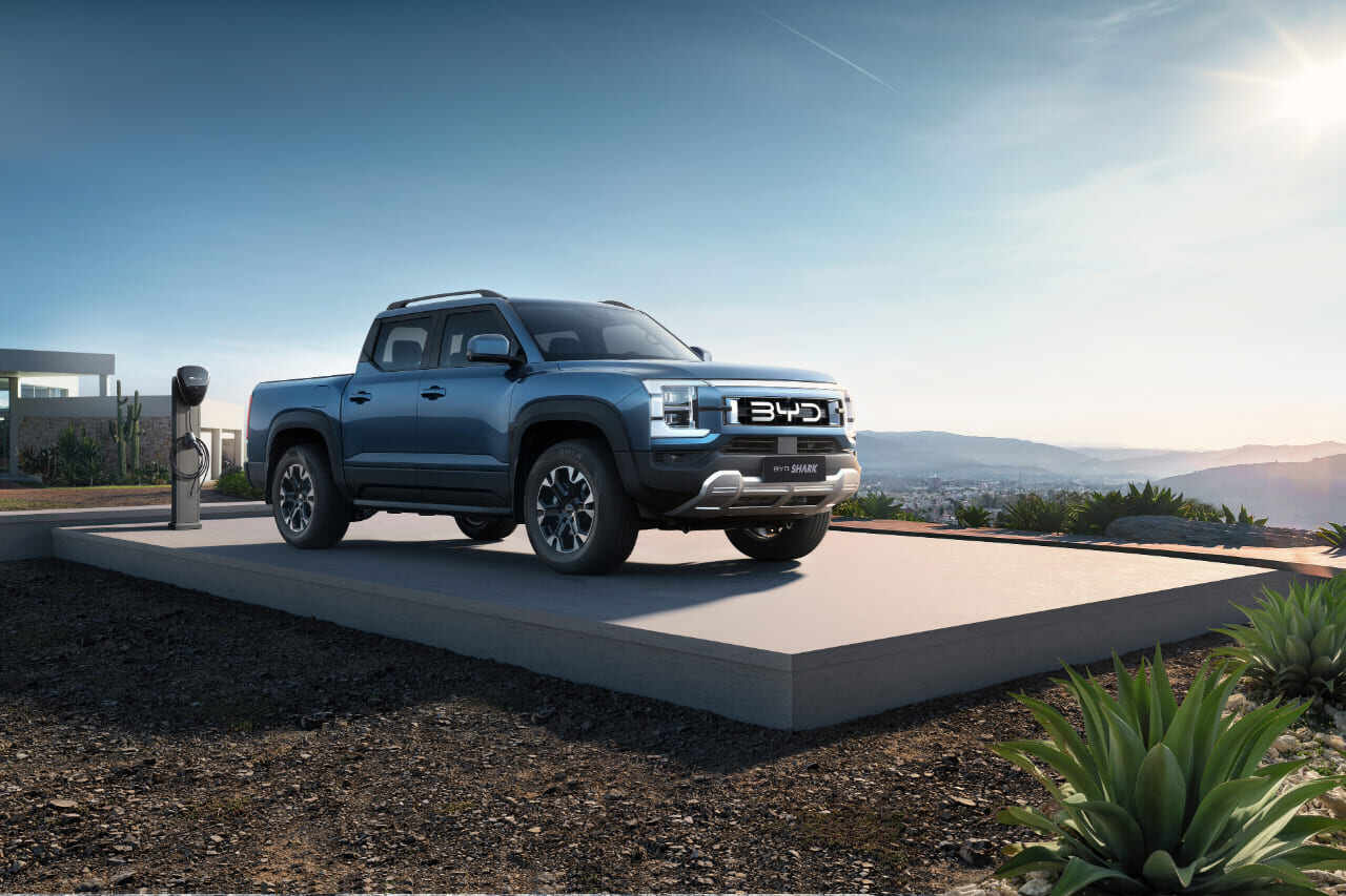 Vista frontal y lateral del pick-up híbrido enchufable BYD SHARK en color azul, estacionado junto a un cargador exterior en un entorno natural y montañoso.